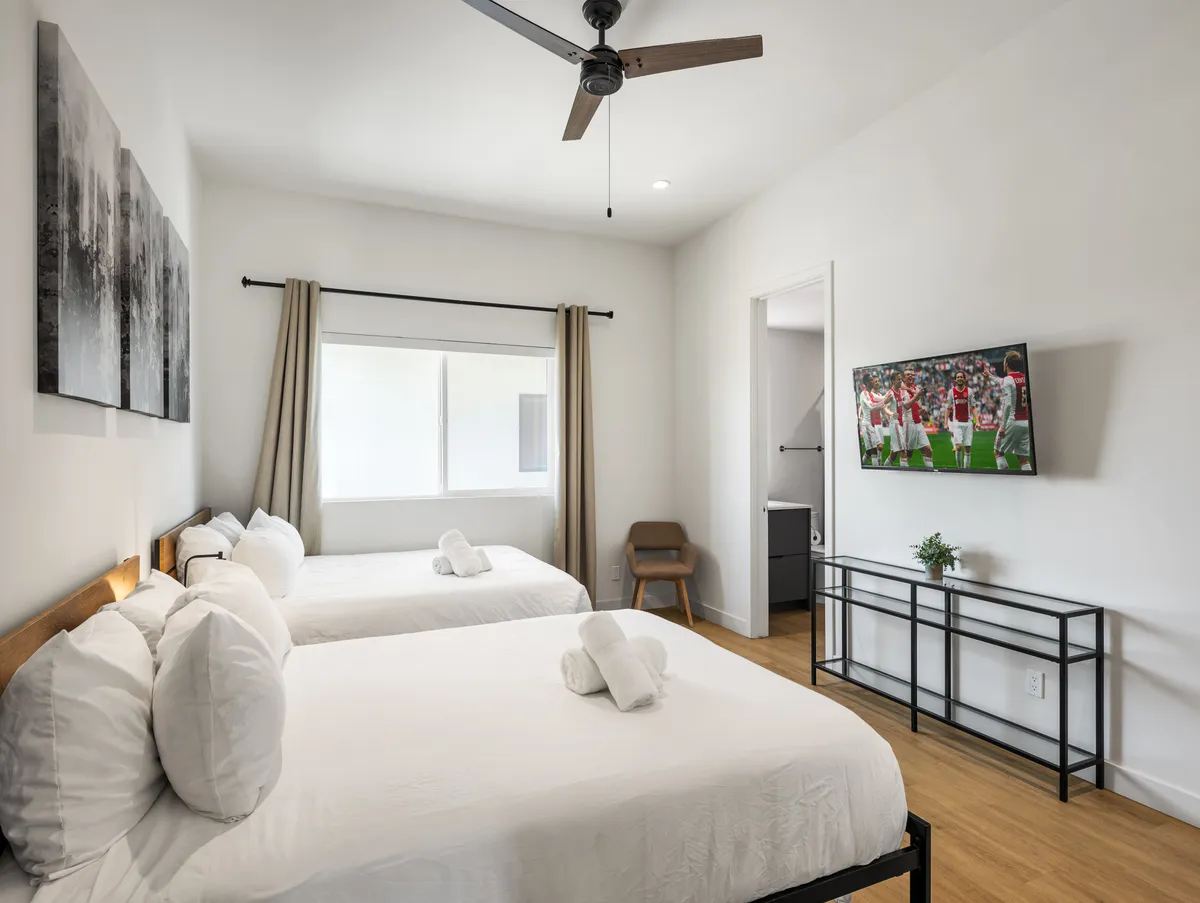 A cozy interior setting featuring two beds with large pillows, a wall-mounted TV displaying a sports event, and vibrant artwork. The room exudes relaxation and comfort, suitable for families or groups.