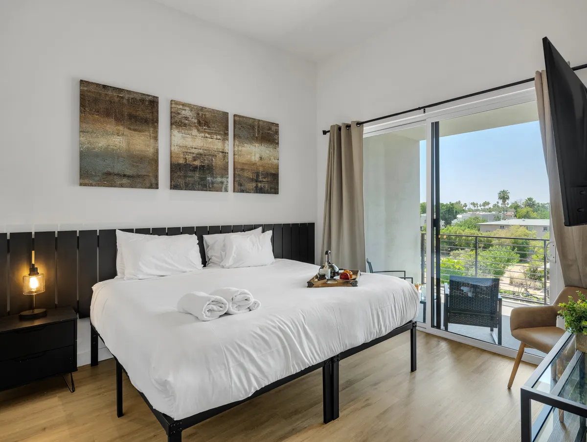 A serene bedroom featuring a bed with crisp white linens, abstract wall art above the headboard, and a balcony visible through sliding glass doors. Natural light streams in, and the room is styled for comfort and tranquility.