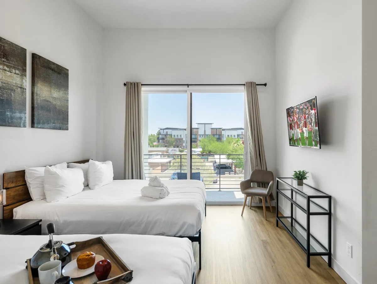 A contemporary bedroom with two beds dressed in white linens, a large window letting in abundant natural light, a wall-mounted TV, a small seating area, and tasteful decorative artwork. The room feels spacious and inviting for groups or families.
