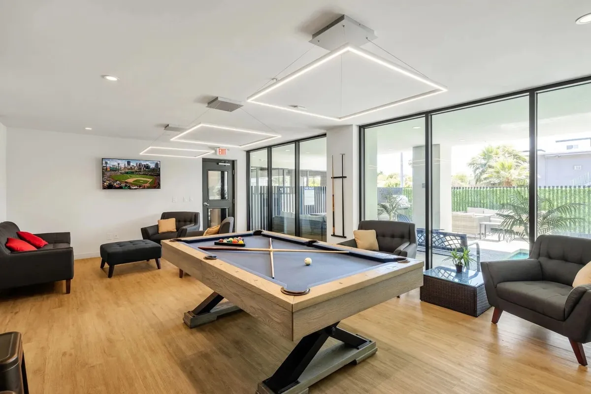 A stylish clubhouse interior featuring a pool table at the center, surrounded by comfortable seating and large windows that fill the space with natural light. The setting encourages friendly games, casual conversation, and group gatherings.