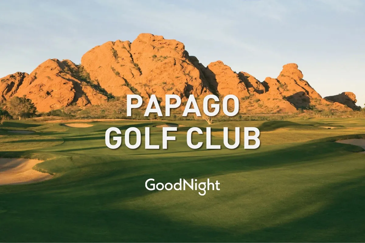 A sweeping view of a lush golf course framed by striking rock formations and a clear blue sky, with the Papago mountains in the background. The scene highlights open green fairways and natural desert landscaping, creating a sense of space and tranquility.